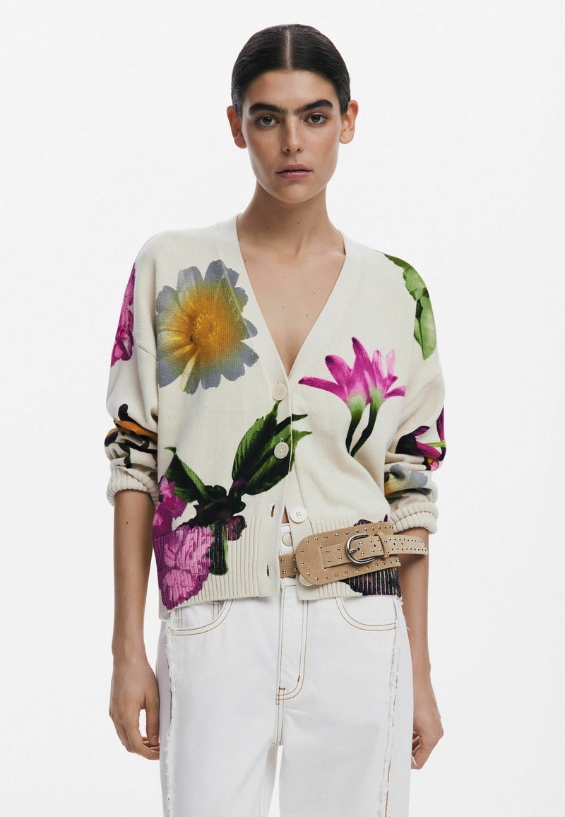 Cardigan fleuri en crème avec des motifs vibrants de violet, jaune et vert. Doté de boutons sur le devant et de poignets côtelés. Porté sur un pantalon blanc.