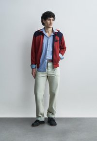 Νεαρός άνδρας με σπαστά μαλλιά, φοράει κόκκινο και ναυτικό τζάκετ, μπλε ριγέ πουκάμισο, ανοιχτό μπεζ παντελόνι, μαύρα παπούτσια, στέκεται μπροστά σε έναν απλό τοίχο.