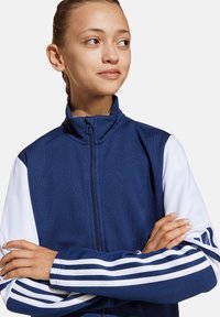 Marineblaue Zip-Jacke mit weißen Ärmeln, ausgestattet mit drei gestreiften Akzenten an den Bündchen und einem hohen Kragen. Der Stoff wirkt glatt und sportlich.