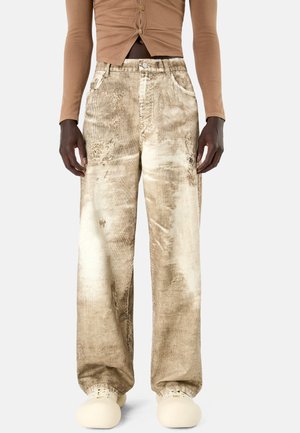 Person trägt beige, mit Säure gewaschene, weit geschnittene Hose, beiges, geknöpftes, kurzes Oberteil und cremefarbene, abgerundete Schuhe, steht vor einem schlichten Hintergrund.