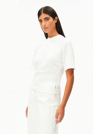 Vrouw met steil donker haar draagt een eenvoudige witte korte mouw top en bijpassende witte rok, poseert tegen een witte achtergrond.