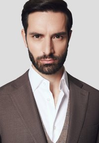 Mężczyzna z przyciętą brodą nosi brązową marynarkę na białej koszuli i wzorzystym kamizelce. Tło jest neutralne szare.