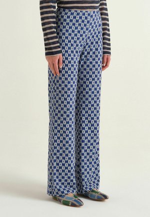 Persona che indossa pantaloni larghi a rombi blu e bianchi, maglia a righe a maniche lunghe e scarpe basse patchwork, in piedi davanti a uno sfondo semplice.