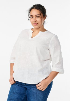 Käharate juustega naine, kes kannab valget tekstuuriga pluusi 3/4 varrukatega ja siniseid teksapükse, poseerib käed taskutes valge tausta ees.