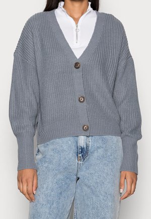 Kvinde iført en grå knap-sat strikket cardigan med V-hals over en hvid lynlåstop og lyseblå jeans med høj talje.