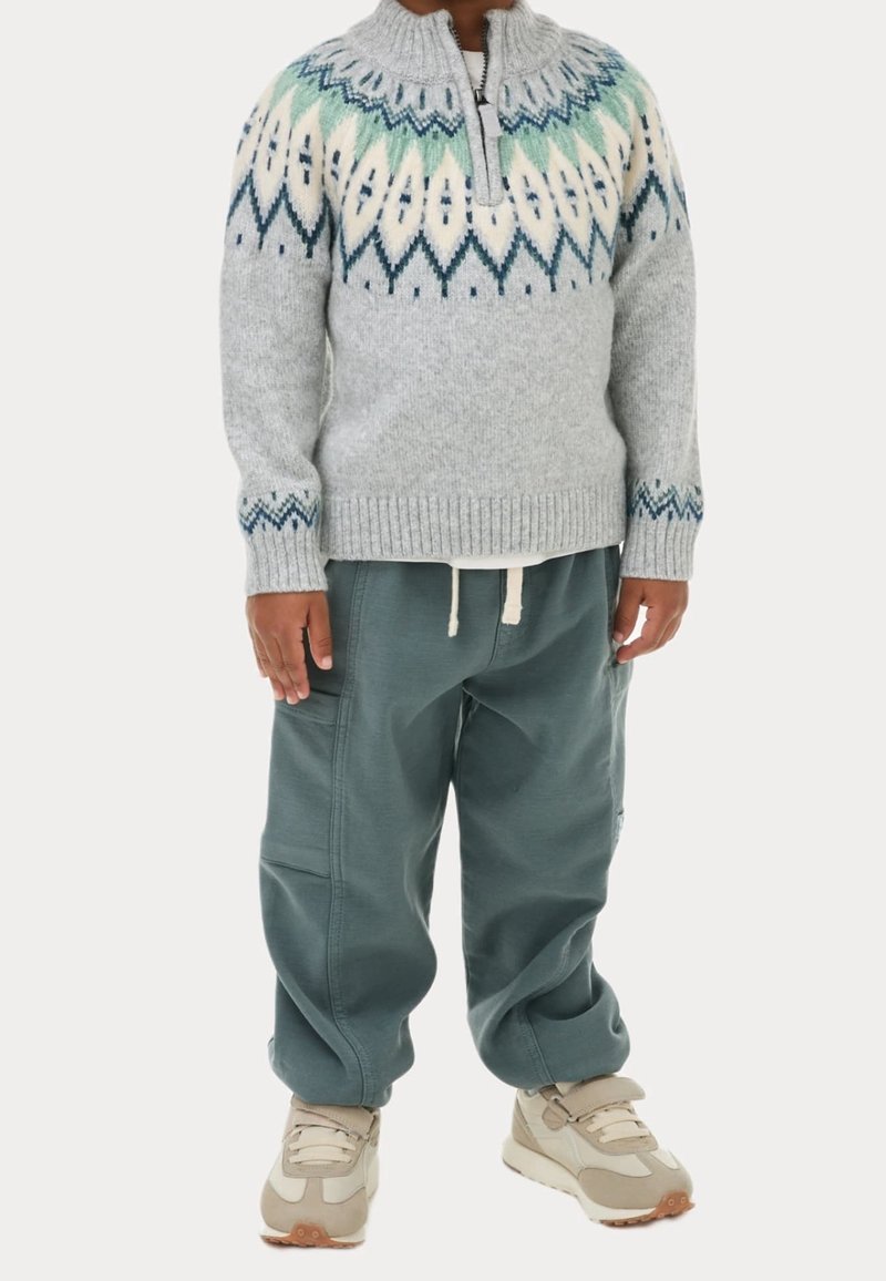 Enfant portant un pull en maille gris à motif, un pantalon cargo ample vert foncé et des baskets beige, debout les mains détendues.
