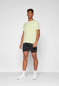 Camiseta de algodão verde-claro com o logotipo "Champion", combinada com calções pretos e ténis brancos. O modelo está em um fundo neutro.
