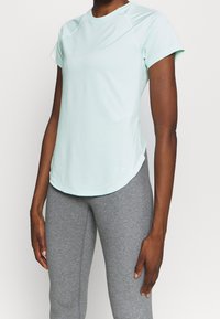 Person som bär en ljusblå kortärmad träningsskjorta och grå tajta leggings, stående framför en enfärgad vit bakgrund.