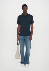 Marine polo skjorte med korte ermer, med en liten logo. Sammen med lyseblå vide jeans og hvite joggesko, holder en beige tote bag.