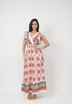 Kobieta stojąca, z ręką na biodrze, ubrana w białą, bezrękawnikową sukienkę maxi z czerwonymi kwiatowymi i paskowymi wzorami oraz beżowe sandały na białym tle.