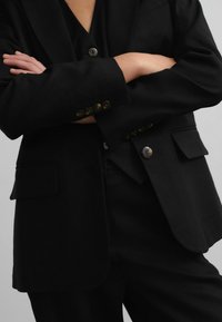 Black tailored blazer with structured shoulders, two flap pockets, and a single button closure. Smooth fabric with a matte finish.