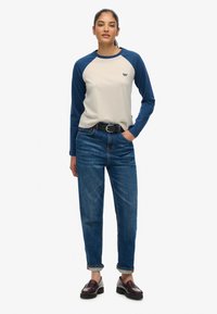 Koszula z długim rękawem, z niebieskimi rękawami raglanowymi i kremowym korpusem, z małym logo. W połączeniu z niebieskimi dżinsami o wysokim stanem i ciemnymi mokasynami.