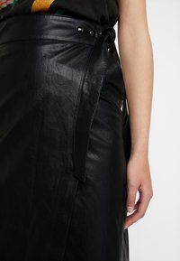 Jupe en cuir noir avec un design à enroulement, dotée d'un lien sur le côté et de boutons noirs brillants. Texture lisse avec des coutures et des plis visibles.