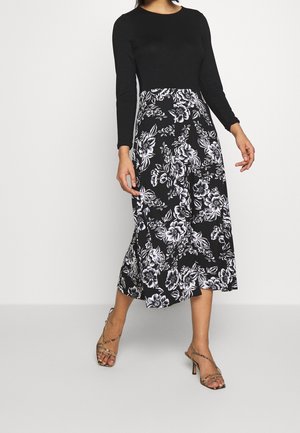 Kobieta ubrana w czarną sukienkę z długim rękawem i białą spódnicę w kwiatowy wzór, nosząca sandały na wysokim obcasie z paskami, stojąca na szarej podłodze.