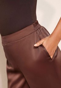 Braune Satin-Hosen mit elastischem Bund. Sie verfügen über eine Seiten- tasche und eine glatte Textur, die durch ein tailliertes Oberteil vor einem neutralen Hintergrund hervorgehoben wird.