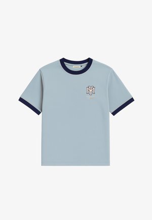 Uomo che indossa una maglietta azzurro chiaro con bordo scuro, bandana blu scuro intorno al collo, pantaloni grigi a righe sottili e cappellino blu scuro con scritta bianca.