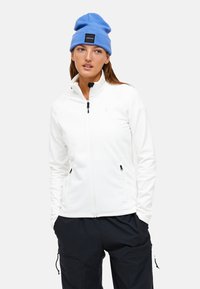 Giacca in pile bianca con colletto alto, zip nera e tasche laterali, abbinata a un berretto di lana blu. Il modello è in piedi su uno sfondo neutro.