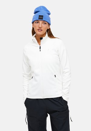 Witte fleece jas met hoge kraag, zwarte rits en zijzakken, gecombineerd met een blauwe gebreide beanie. Model staat tegen een neutrale achtergrond.