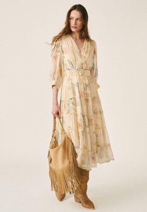 Mujer con un vestido largo y pálido con estampado floral y cinturón, sujetando un bolso marrón con flecos, y llevando botas de vaquero marrones a juego sobre un fondo liso.