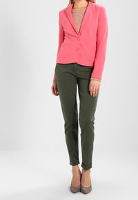 Roze getextureerde blazer met twee zijzakken, gedragen over een beige gebreid sweater. Gecombineerd met olijfgroene broek en grijze hoge hakken.