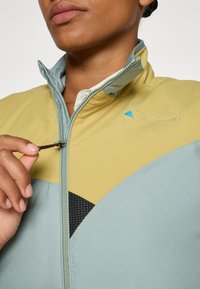 Veste zippée légère avec un design bicolore en vert pâle et jaune. Panneau en mesh noir texturé et détail de marque discret.