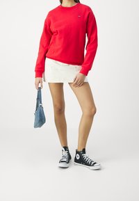 Femme portant une chemise rouge à manches longues, une mini-jupe blanche, des baskets montantes noires, tenant un petit sac à main bleu sur un fond uni.