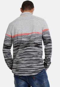 Chemise grise à manches longues avec une rayure rouge horizontale, présentant un motif texturé dans des teintes plus sombres, avec des poignets à boutons et un col.