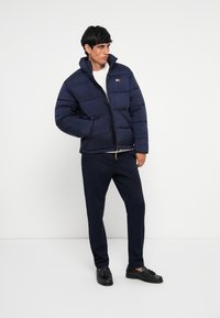 Marineblauer Pufferjacke mit hohem Kragen, gestepptem Design und Frontreißverschluss. Kombiniert mit marineblauen Hosen und schwarzen Slippern. Weißer Pullover darunter.
