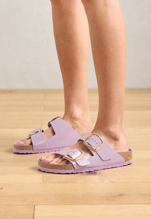 Asmuo dėvi pastelinės violetinės spalvos šlepetes su dviem plačiais diržais ir auksiniais užsegimais ant medinio grindų paviršiaus.