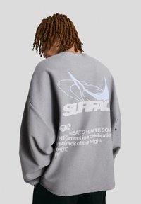 Šedá oversize mikina s grafikou a nápisem "SURFACE" v bílé barvě. Látka vypadá měkká s texturovaným povrchem.