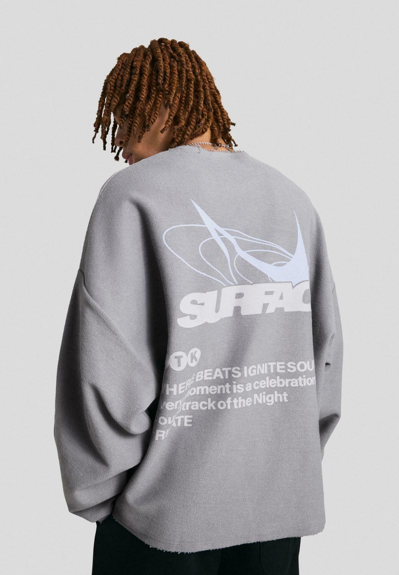 Šedá oversize mikina s grafikou a nápisem "SURFACE" v bílé barvě. Látka vypadá měkká s texturovaným povrchem.