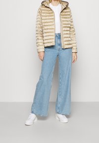 Beige péřová bunda s prošívaným designem, kapucí, na zip, spárovaná se světle modrými širokými džíny a bílými teniskami.