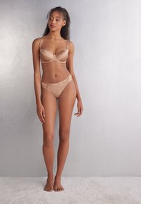 Svetle hnědé saténové podprsenka a odpovídající kalhotky, které mají nastavitelné ramínka a hladký povrch, předváděné na modelce na neutrálním pozadí.