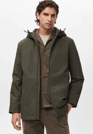 Chaqueta verde oliva resistente al agua con capucha, cierre de cremallera y bolsillos laterales, superpuesta sobre un suéter de punto beige y una camisa marrón.