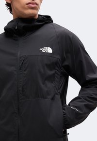 Άνδρας που φοράει μαύρο μπουφάν The North Face με τσεπάκι στο στήθος με φερμουάρ και κουκούλα, κοιτάζει ελαφρώς προς τα αριστερά σε καθαρό ανοιχτό φόντο.