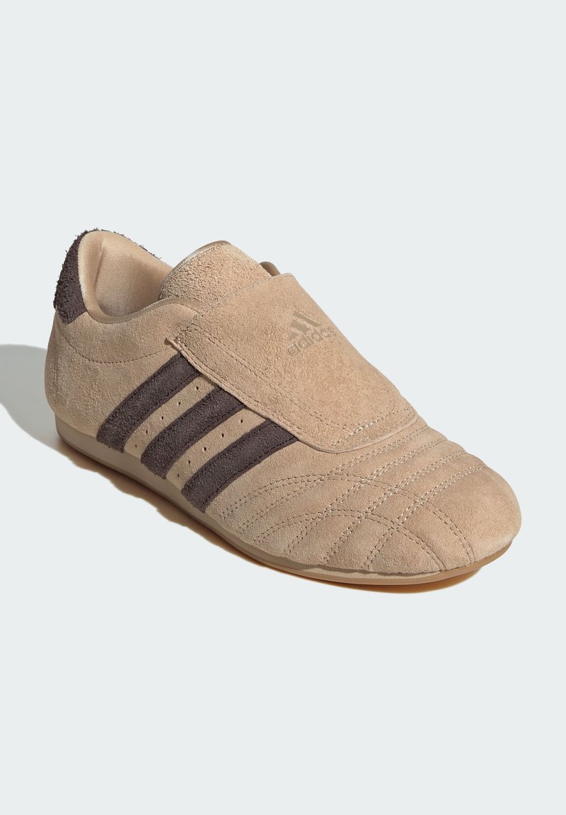 Zapatilla deportiva de ante en color beige claro con franjas marrón oscuro. Presenta una puntera redondeada, diseño sin cordones y detalles texturizados en la parte superior.