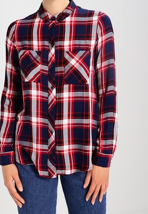 Nainen pukeutuneena punaiseen, laivastonsiniseen ja valkoiseen ruudulliseen nappipaitaan, jossa on kaksi rintataskua, ja sinisiin farkkuihin, seisoo kädet rentoina sivuilla.