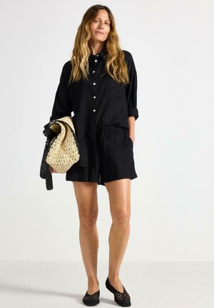 Camicetta nera a maniche corte con chiusura a bottoni, pantaloni corti con vita elastica, scarpe nere slip-on e una borsa a tracolla intrecciata con dettagli a righe.