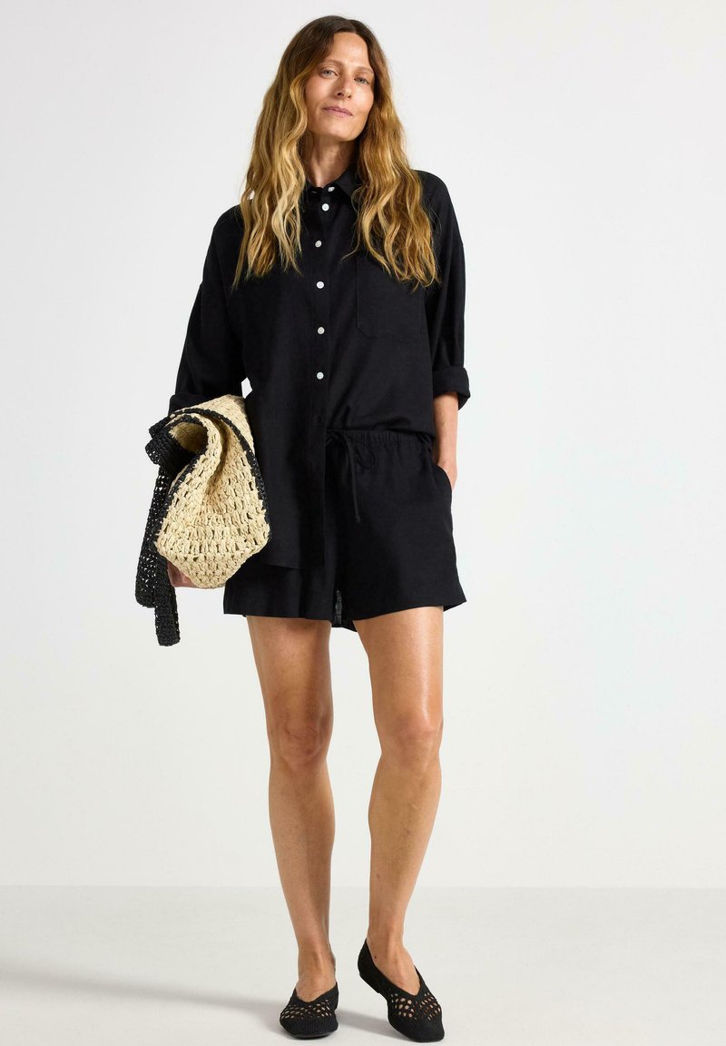 Camicetta nera a maniche corte con chiusura a bottoni, pantaloni corti con vita elastica, scarpe nere slip-on e una borsa a tracolla intrecciata con dettagli a righe.