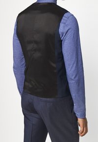 Svart satinryggväst med en figurnära design, använd över en blå strukturerad skjorta. Observera den mjuka tygövergången och den mörka färgkombinationen.