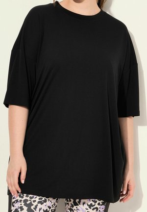 Personne portant une chemise noire ample à manches courtes associée à des leggings imprimés léopard blancs et noirs, debout devant un fond clair.