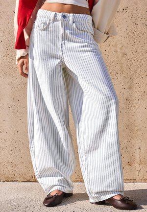 Wide-leg white pants with navy vertical stripes worn with brown ballet flats and a red sweater draped over the shoulders.