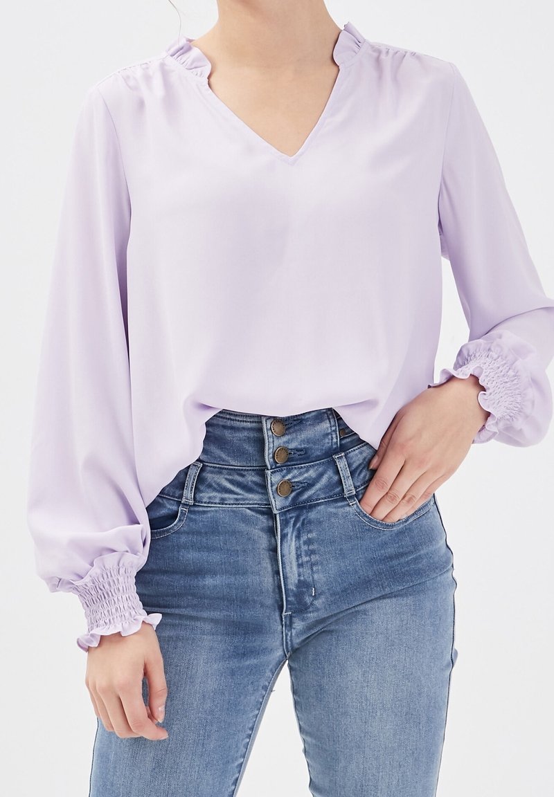 Blusa lavanda con scollo a V, colletto arricciato e maniche lunghe a sbuffo. Abbinata a jeans in denim a vita alta con dettagli sui bottoni.