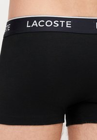 Czarne bawełniane bokserki z granatowym pasem, na którym widnieje białe logo "LACOSTE". Gładka tekstura, dopasowany krój i brak widocznych wzorów.