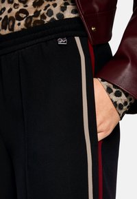 Black pants featuring a textured fabric, side panel stripes in beige and red, elastic waistband, and a small logo tag near the pocket.
