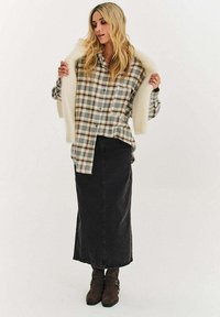 Károvaná flanelová košeľa v prírodných tónoch s voľným strihom, vrstvená pod krémovou šálovou kožušinkou, doplnená dlhou tmavou rifľovou sukňou a hnedými čižmami.