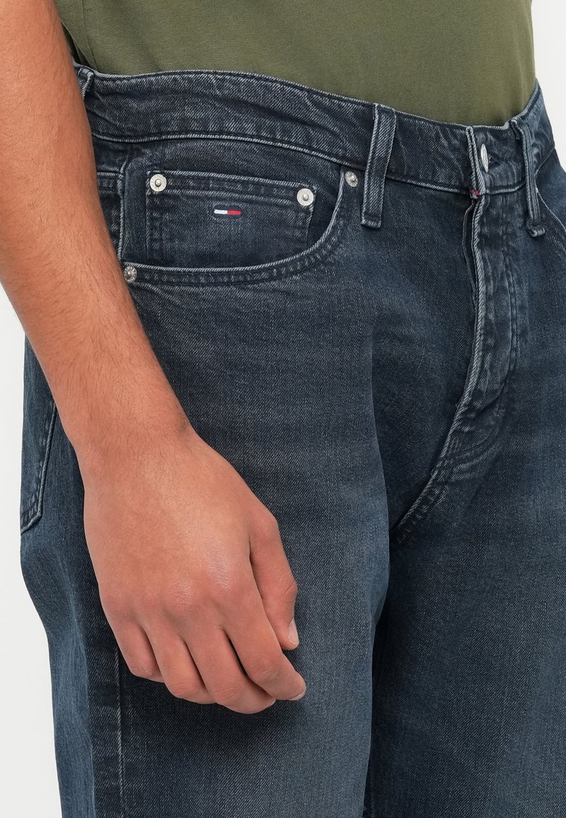 Dark denim jeans with a regular fit, featuring metal hardware and a small logo patch on the front pocket. Smooth texture, classic design.