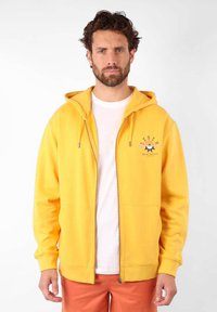 Hoodie zippé jaune en tissu doux, avec une poche avant, une capuche avec cordons de serrage, et un imprimé graphique sur la poitrine.