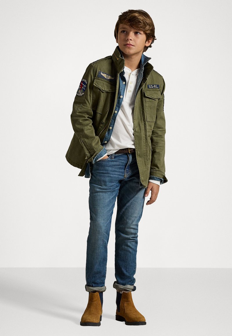 Olivgrön militärstil-jacka med flera fickor och lappar, i kombination med en vit skjorta, blå jeans och bruna ankelskor.