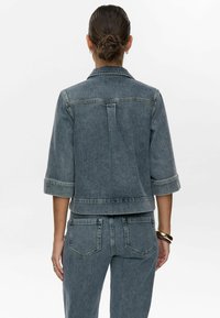 Temno denimjakna skrajšane dolžine z zavihanimi rokavi, ki ima strukturni ovratnik, detajl zadnjega jermena in zadnji pregib. V kombinaciji z usklajenimi jeans hlačami.